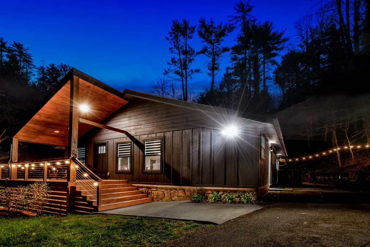 The warmly illuminated porch of the cabin, featuring string lights, wooden facade, and steps leading up to the entrance. The space is inviting and set up for gatherings, with a tranquil wooded backdrop. The mood is welcoming and perfect for social evenings or starting a new day together.