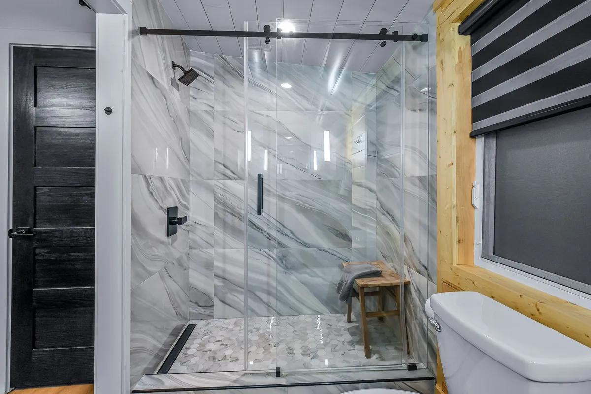 A tranquil bathroom featuring a glass shower enclosure, elegant marble walls, a wooden stool, and modern fixtures. The space is bright, fresh, and spa-like, ideal for rejuvenating showers and self-care routines.