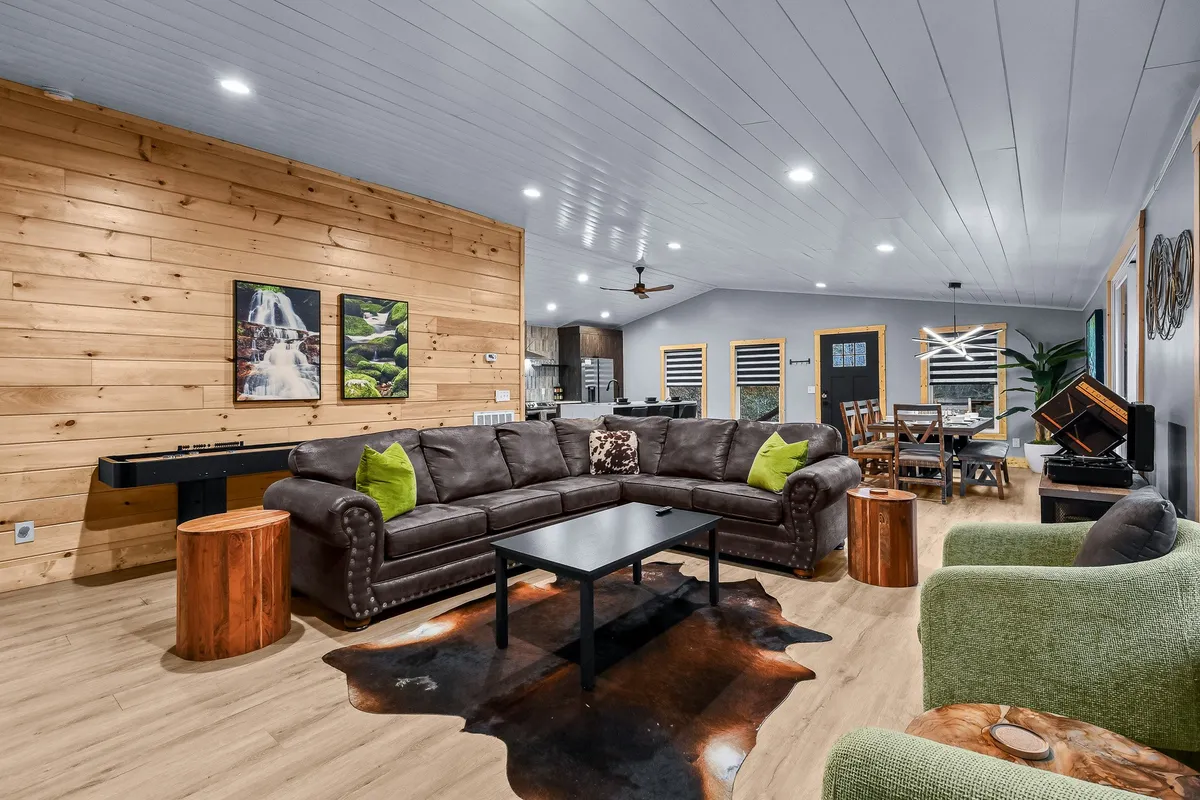 A spacious, open-concept living room featuring a large brown sectional couch, a central coffee table, and a wooden wall adorned with artwork. The setting is inviting, designed for relaxation, socializing, and group activities.