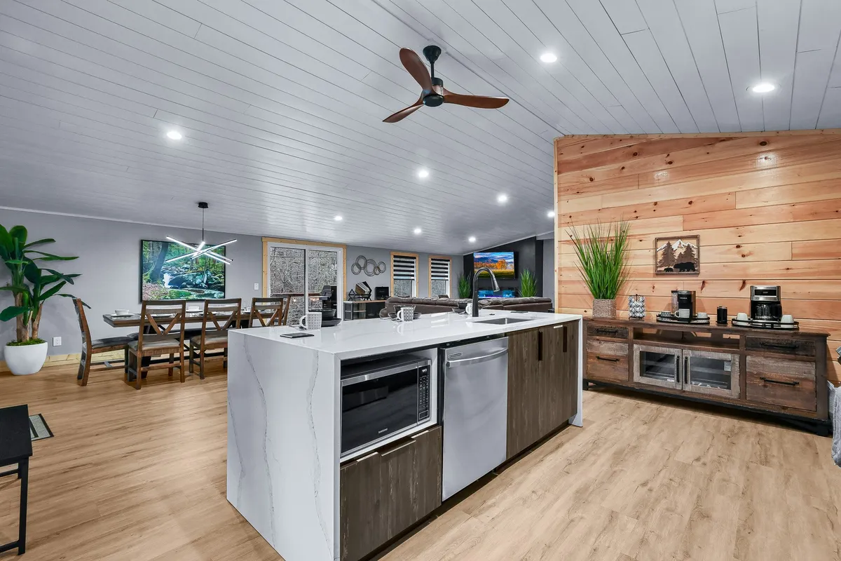 A contemporary, open-plan kitchen and dining area featuring a spacious kitchen island, sleek stainless appliances, a wooden accent wall, and large windows that flood the space with natural light. The dining table is visible and inviting, with a clear flow into the living area.