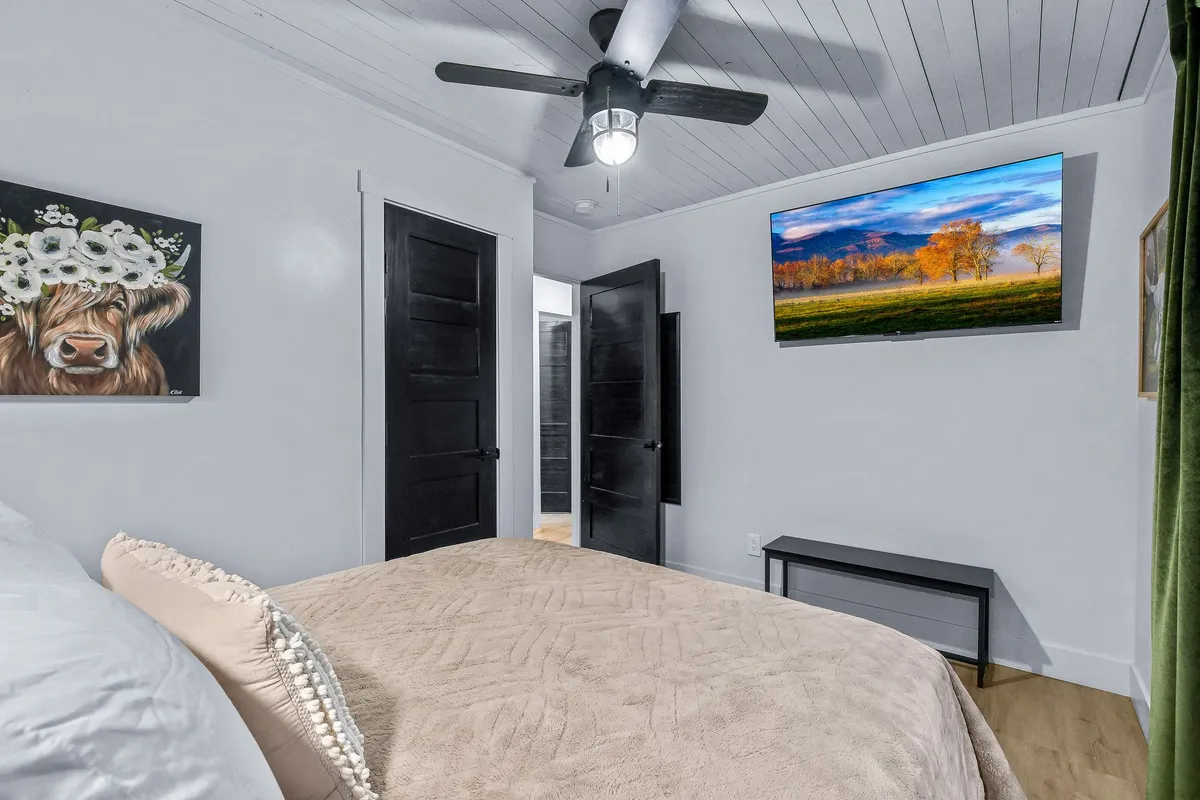 A peaceful bedroom featuring a plush bed with a soft blanket, artwork of a cow on the wall, and a flat-screen TV displaying a calming landscape. Sunlight pours in from open doors, creating a serene and restful atmosphere ideal for relaxation and unwinding.