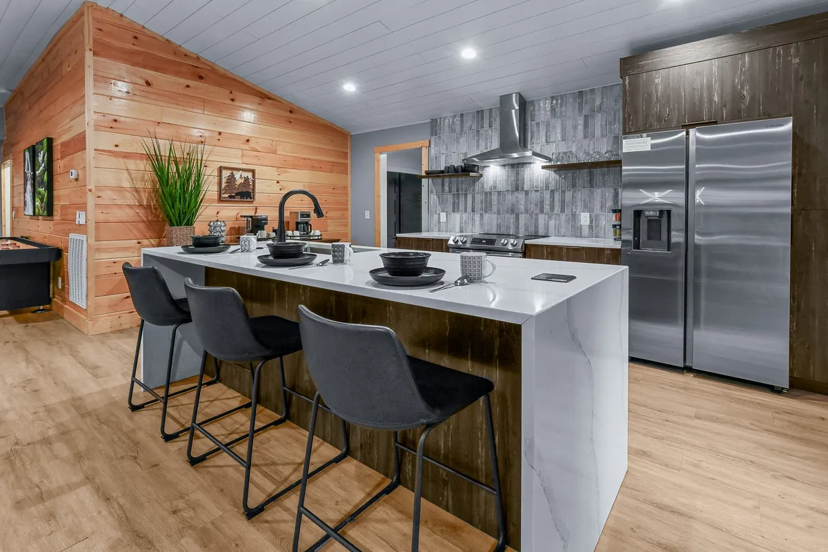 A stylish kitchen with a central island, modern stainless steel appliances, wooden wall accents, and dark dishware neatly arranged. The space exudes warmth and readiness for cooking or entertaining.