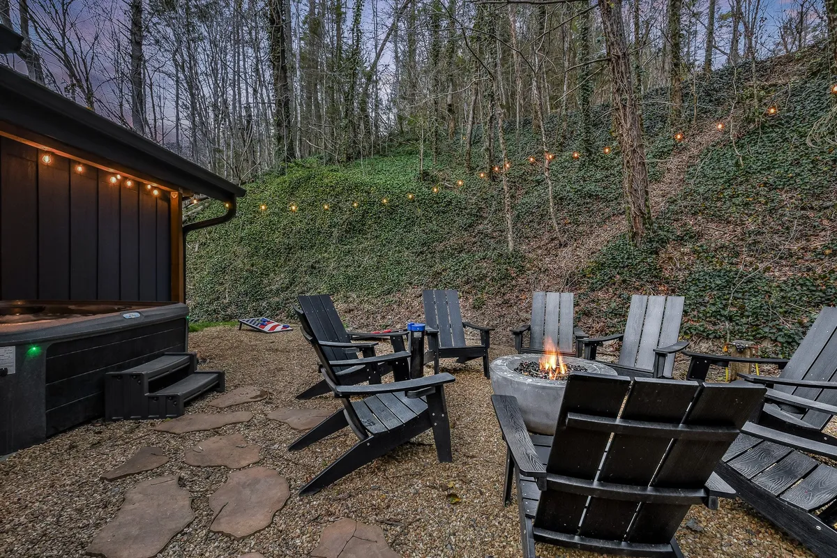 A cozy outdoor area featuring a fire pit surrounded by adirondack chairs, a bubbling hot tub, and string lights overhead. The atmosphere is relaxed, inviting guests to gather around the fire or soak under the stars.