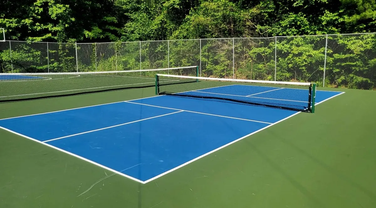 A well-kept tennis court surrounded by lush greenery, featuring a prominent net and crisp court lines under a sunny sky. The area is clearly set up for group activities such as playing tennis, practicing serves, or simply socializing outside.