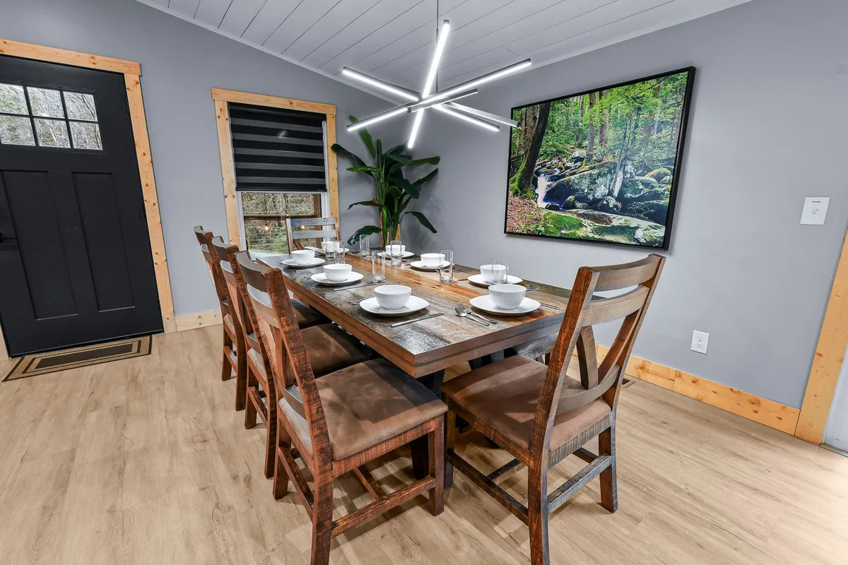 A generously sized dining table with a modern light fixture above, nature-inspired artwork, and a leafy indoor plant, creating a cozy and welcoming atmosphere suitable for group meals and celebrations.