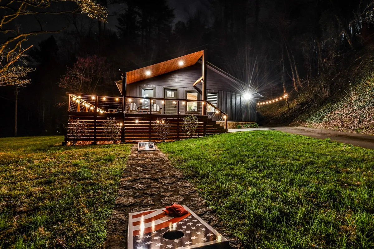 A modern cabin exterior is adorned with string lights and features cornhole boards set up in the foreground, inviting friendly outdoor games. The mood is festive and relaxed, perfect for group gatherings.