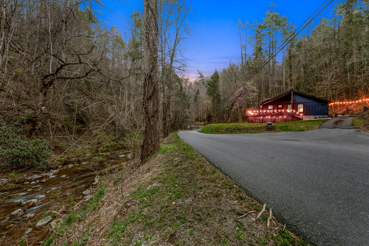 A serene view of the blue house nestled beside a winding road and a peaceful creek, illuminated by warm string lights. The house sits in a lush, wooded setting, perfectly blending with the surrounding nature. The porch and road offer vantage points for taking in the tranquil scenery.